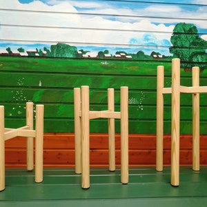 Può includere: Quattro portavasi in legno di diverse altezze, realizzati in legno naturale con finitura marrone chiaro. I portavasi sono disposti in fila su una superficie verde, con un murale dipinto di una scena di campagna sullo sfondo.