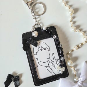 May include: Black ID badge holder with a silver keychain, a black bow, and a heart charm. The holder features a black and white illustration of a person making a peace sign. A pearl necklace and white bow are also present.
