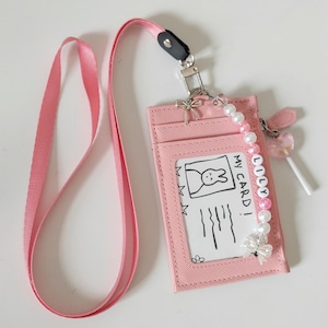 Puede incluir: Tarjetero rosa con cordón a juego. El tarjetero tiene una ventana transparente con una ilustración de conejito y el texto "MY CARD!". Un charm de cuentas con el nombre "LILY" y un charm de piruleta están adjuntos.
