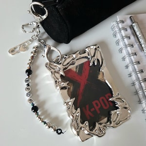Puede incluir: Llavero de metal plateado con un colgante rectangular. El colgante tiene un fondo negro con un diseño de 'X' rojo y las palabras 'K-POP'. Un dije de cuentas con las letras 'KPOP' está adjunto.