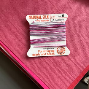 Może przedstawiać: Szpula naturalnej nici jedwabnej, głównie różowej i białej, z napisem "NATURAL SILK" i "For stringing pearls and beads". Nić ma 2 metry długości i średnicę 0,70 mm. Szpula leży na różowej, teksturowanej powierzchni.