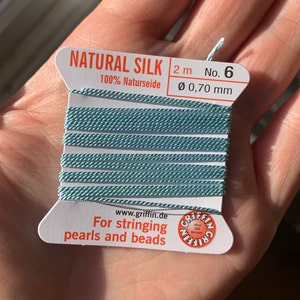Può includere: Un rocchetto di 2 metri di filo di seta naturale blu chiaro, 100% seta naturale, con un diametro di 0,70 mm. Il rocchetto è etichettato "NATURAL SILK 2 m No. 6 100% Naturseide Ø 0,70 mm" e "Per infilare perle e perline".