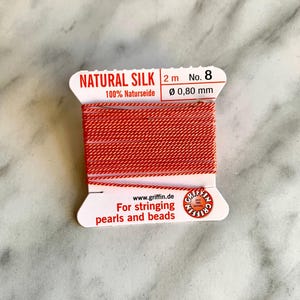 Peut inclure: Une bobine de fil de soie naturelle rouge pour enfiler des perles et des perles. La bobine est étiquetée "NATURAL SILK 2 m No. 8 100% Naturseide Ø 0,80 mm" et "For stringing pearls and beads".
