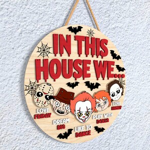 May include: A circular wooden sign with the text "IN THIS HOUSE WE" in red, and the phrases "LOVE FRIDAY", "DREAM BIG", "LIKE TO DANCE", "PLAY WITH DOLLS", and "LOVE FAMILY". The sign features cartoon horror characters and black bat silhouettes.