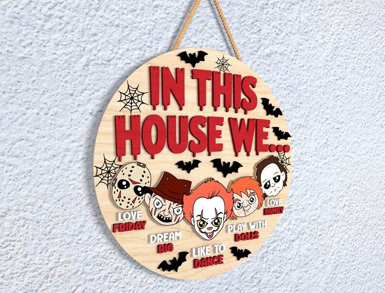 May include: A circular wooden sign with the text "IN THIS HOUSE WE" in red, and the phrases "LOVE FRIDAY", "DREAM BIG", "LIKE TO DANCE", "PLAY WITH DOLLS", and "LOVE FAMILY". The sign features cartoon horror characters and black bat silhouettes.