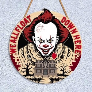 May include: Round wooden sign featuring a menacing clown with red hair and makeup, a red balloon, and a haunted house. The sign reads "WE ALL FLOAT DOWN HERE" in red lettering, perfect for Halloween decor.
