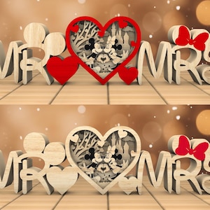 Può includere: Taglio in legno di "Mr." e "Mrs." con un design a forma di cuore al centro. Il cuore presenta una silhouette di Topolino e Minnie Mouse all'interno di un castello.