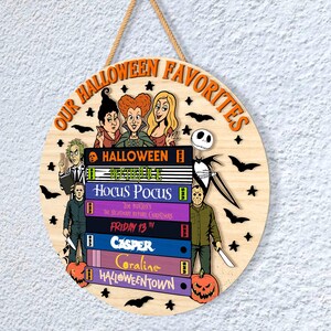 May include: Circular wooden Halloween sign with the text "OUR HALLOWEEN FAVORITES" in orange. The sign features illustrations of Halloween characters and movie titles like "Hocus Pocus" and "Friday the 13th". Includes a jute rope for hanging.