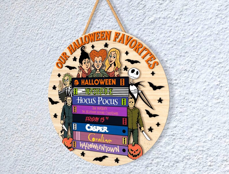 May include: Circular wooden Halloween sign with the text "OUR HALLOWEEN FAVORITES" in orange. The sign features illustrations of Halloween characters and movie titles like "Hocus Pocus" and "Friday the 13th". Includes a jute rope for hanging.