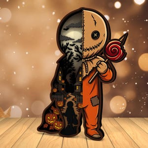Może przedstawiać: Wycięcie z drewna o tematyce Halloween, przedstawiające postać w pomarańczowym kombinezonie trzymającą lizaka. Głowa postaci jest podzielona, z jednej strony pokazującą zszytą maskę, a z drugiej upiorny krajobraz z domem, dyniami i sylwetką dziecka.