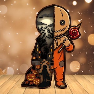 Może przedstawiać: Wycięcie z drewna o tematyce Halloween. Postać ma na sobie pomarańczowy kombinezon i maskę z worka jutowego, trzymając lizaka. Lewa strona przedstawia upiorną scenę z domem, dyniami i sylwetką.