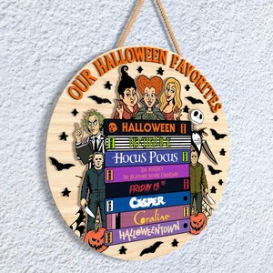 May include: Circular wooden Halloween sign with the text "OUR HALLOWEEN FAVORITES". The sign features illustrations of characters from classic Halloween films, including Beetlejuice, Michael Myers, and the Sanderson sisters.