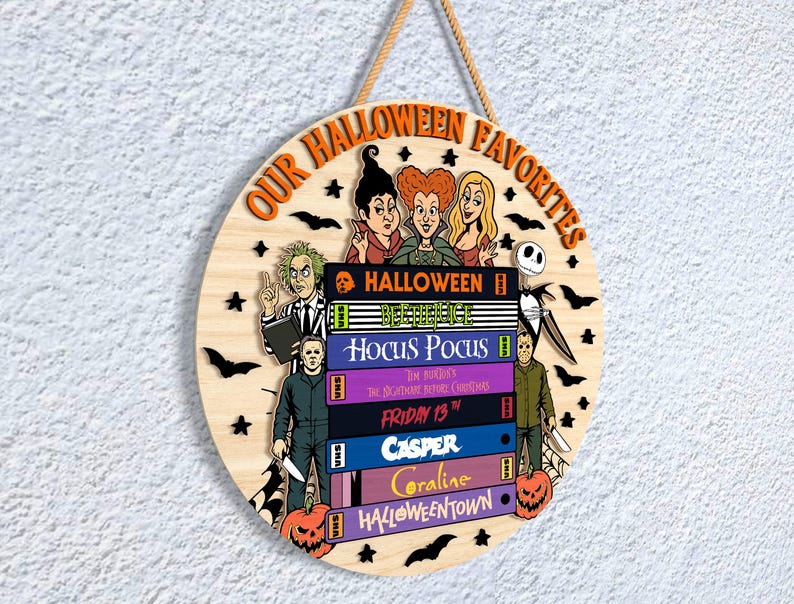 May include: Circular wooden Halloween sign with the text "OUR HALLOWEEN FAVORITES". The sign features illustrations of characters from classic Halloween films, including Beetlejuice, Michael Myers, and the Sanderson sisters.