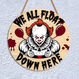 May include: Round wooden sign featuring a menacing clown with red hair and makeup, holding a paper boat. The sign reads "WE ALL FLOAT DOWN HERE" in black letters, with red balloon and blood splatter accents. A rope is attached for hanging.