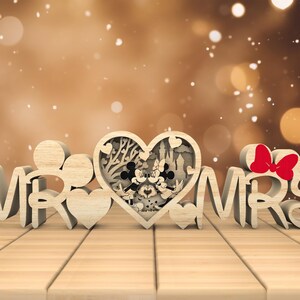 May include: Wooden "Mr. & Mrs." sign with a heart-shaped cutout featuring a silhouette of Mickey and Minnie Mouse. The heart is surrounded by a floral design.