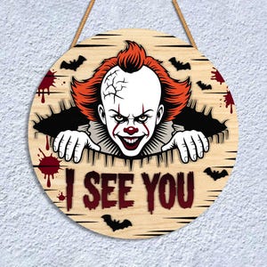 May include: Round wooden sign featuring a scary clown with red hair and makeup, emerging from a hole. The sign includes the text "I SEE YOU", bats, and blood splatter designs. A rope is attached for hanging.
