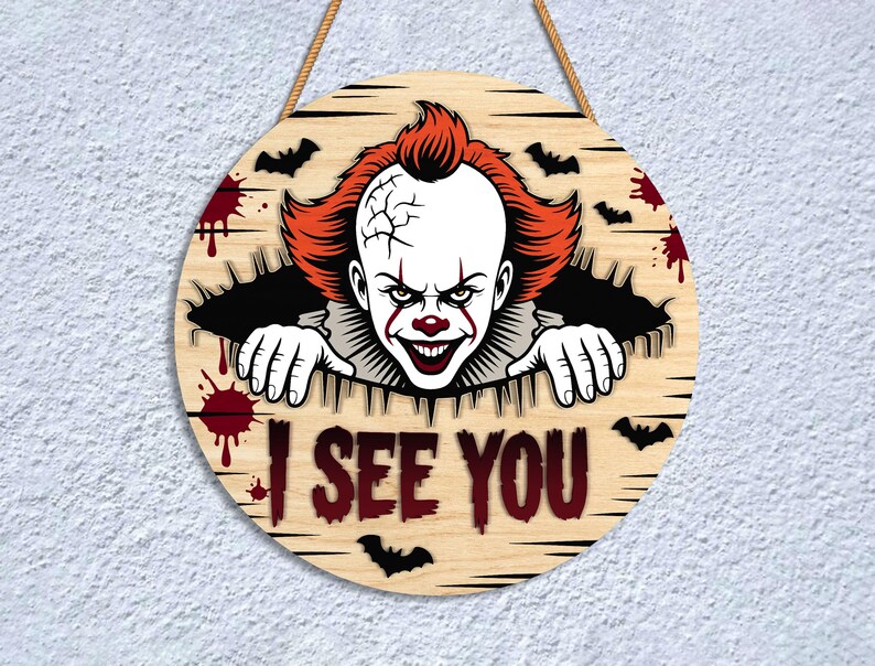 May include: Round wooden sign featuring a scary clown with red hair and makeup, emerging from a hole. The sign includes the text "I SEE YOU", bats, and blood splatter designs. A rope is attached for hanging.