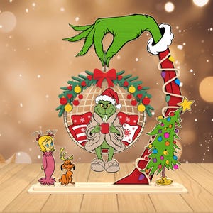 Pode incluir: Decoração de Natal em madeira com o Grinch, Cindy Lou Who e Max. O Grinch, com gorro de Papai Noel e roupão, segura uma caneca. Uma mão verde paira sobre uma coroa. Uma pequena árvore de Natal também está incluída.