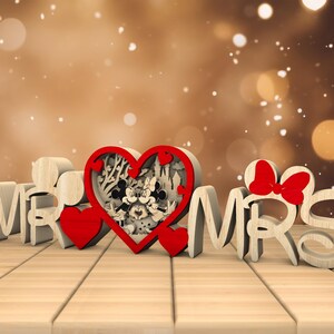 May include: Wooden "Mr. & Mrs." sign with a red heart in the centre featuring a Mickey Mouse and Minnie Mouse design. The heart is surrounded by a decorative floral pattern.