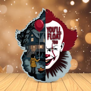 Puede incluir: Decoración de madera en capas con un diseño dividido. Un lado representa un payaso siniestro con el texto "YOU'LL FLOAT TOO". El otro lado muestra a un niño con un abrigo amarillo sosteniendo un globo rojo frente a una casa embrujada.
