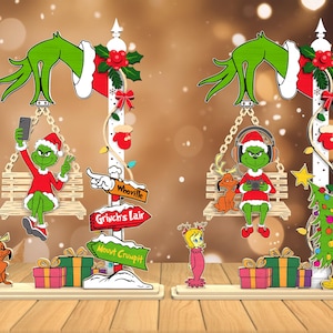 Pode incluir: Decorações de Natal com tema Grinch. Dois Grinch estão em balanços, um tirando uma selfie e o outro usando fones de ouvido. Cada um tem um poste de sinalização com sinais direcionais. Presentes e uma árvore de Natal também estão incluídos.