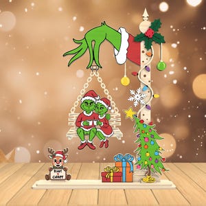 May include: A whimsical Christmas decoration featuring the Grinch and Cindy Lou Who on a swing, with a Christmas tree, presents, and a reindeer holding a sign that says "Ruddy Carrot". The Grinch's hand holds the swing, with festive lights and ornaments.