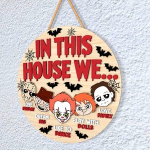 May include: Circular wooden sign with the text "IN THIS HOUSE WE..." in red, featuring horror movie character illustrations. Includes the phrases "LOVE FRIDAY", "DREAM BIG", "LIKE TO DANCE", "PLAY WITH DOLLS", and "LOVE FAMILY". Decorated with bats and spiderwebs.