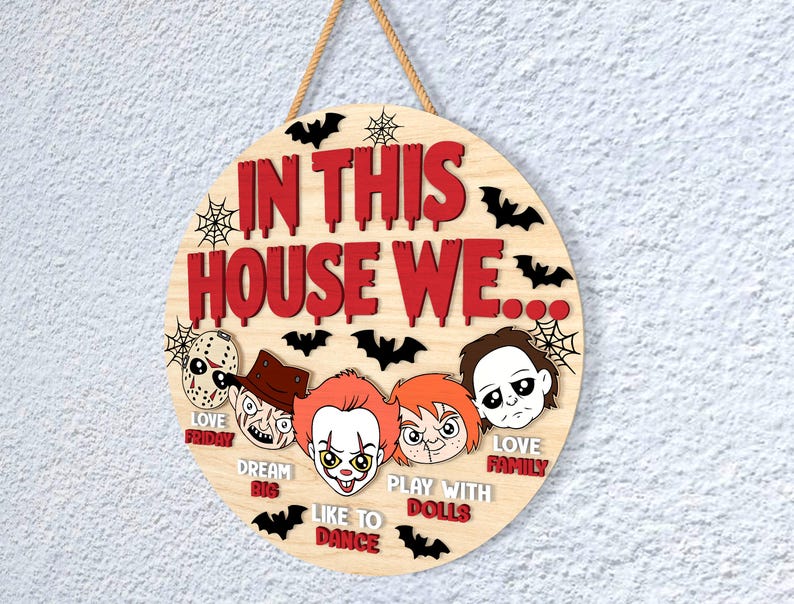 May include: Circular wooden sign with the text "IN THIS HOUSE WE..." in red, featuring horror movie character illustrations. Includes the phrases "LOVE FRIDAY", "DREAM BIG", "LIKE TO DANCE", "PLAY WITH DOLLS", and "LOVE FAMILY". Decorated with bats and spiderwebs.