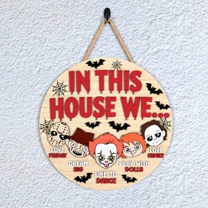 May include: Circular wooden sign with the text "IN THIS HOUSE WE..." in red. The sign features cartoon images of horror movie characters and bats. The sign is suspended by a rope.
