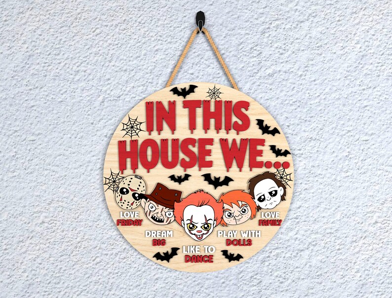 May include: Circular wooden sign with the text "IN THIS HOUSE WE..." in red. The sign features cartoon images of horror movie characters and bats. The sign is suspended by a rope.