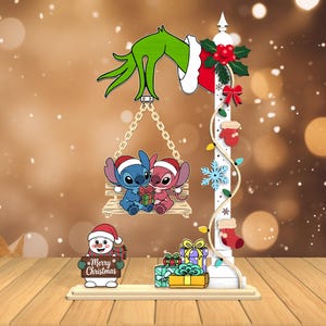 Peut inclure: Décoration de Noël avec Stitch et Angel sur une balançoire, tenue par une main du Grinch. Un bonhomme de neige tient une pancarte "Merry Christmas". Des cadeaux et un poteau décoré avec des lumières et des ornements complètent la scène.