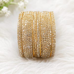 May include: A set of four gold-toned bangles, each featuring a dense, uniform surface of small, sparkling clear crystals. The bracelets are arranged side-by-side, showcasing a continuous, reflective metallic finish and a high-shine, textured appearance.