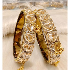 May include: Two gold-toned metal bangles featuring a repeating pattern of heart-shaped champagne-coloured crystals bordered by small white beads. Each bangle is accented with dangling gold-toned metal charms and a smooth, reflective inner surface.