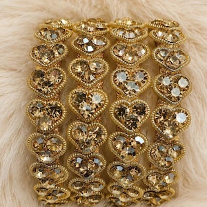 May include: A set of four gold-toned bangles arranged side by side. Each bangle features a repeating pattern of heart-shaped settings, with each heart containing a cluster of small, clear, faceted crystals that reflect light across the surface.