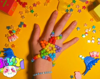 Puede incluir: Una mano lleva una pulsera de cuentas con forma de estrella arcoíris. La pulsera está hecha de cuentas de plástico de colores y se lleva en la muñeca y los dedos. El fondo es amarillo con estrellas arcoíris dispersas.