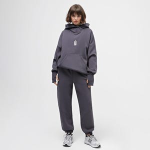May include: A charcoal gray hoodie and matching sweatpants set. The hoodie features a front pocket and a white graphic on the chest. The sweatpants have a relaxed fit. The model is wearing silver and white sneakers.
