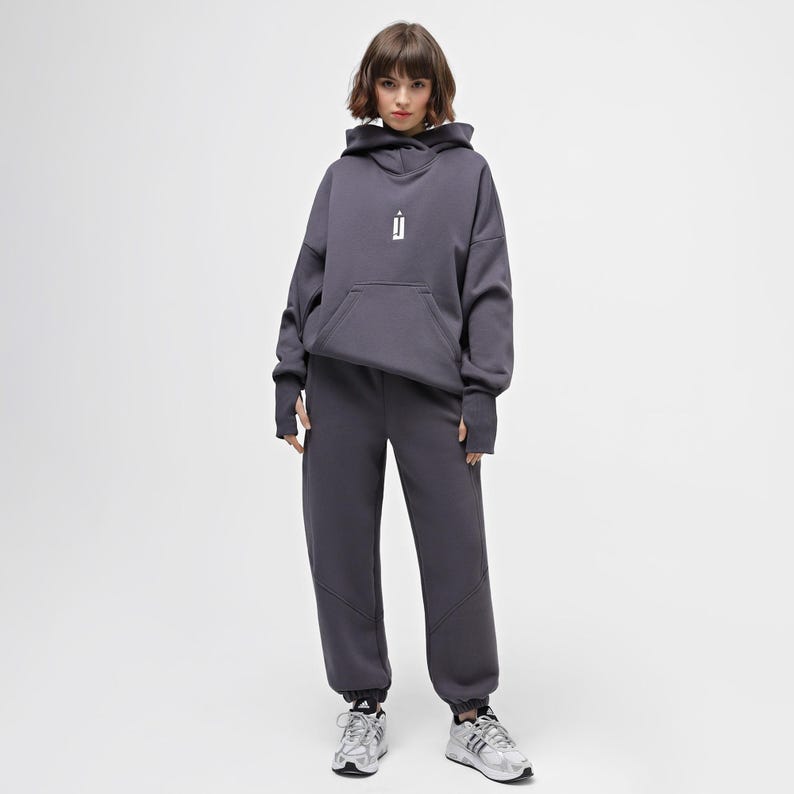 May include: A charcoal gray hoodie and matching sweatpants set. The hoodie features a front pocket and a white graphic on the chest. The sweatpants have a relaxed fit. The model is wearing silver and white sneakers.