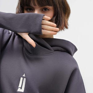 May include: Charcoal gray hoodie with a white logo on the front. The hoodie features a hood and ribbed cuffs. The image shows a close-up of the hoodie, with a person's hand covering their mouth.
