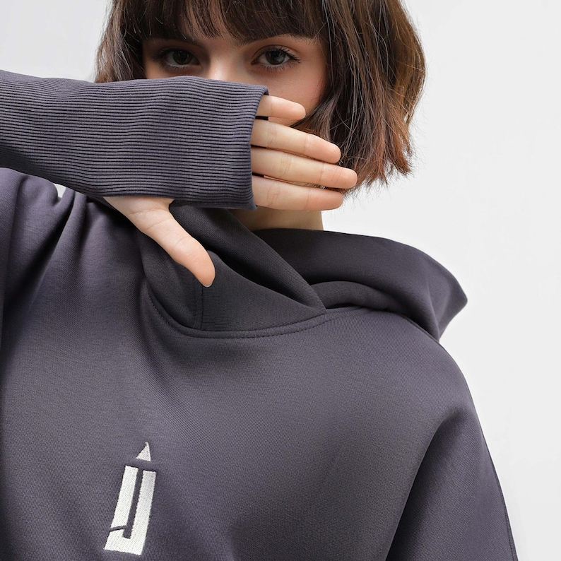 May include: Charcoal gray hoodie with a white logo on the front. The hoodie features a hood and ribbed cuffs. The image shows a close-up of the hoodie, with a person's hand covering their mouth.