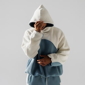May include: A two-tone hoodie and matching sweatpants set. The hoodie is white with a blue panel across the chest and a large hood. The sweatpants are blue with white accents. The person in the image is wearing the hoodie and sweatpants.