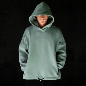 May include: A sage green hoodie with a drawstring at the waist. The hooded sweatshirt has a relaxed fit and long sleeves. The person in the image is wearing black pants. The background is black.