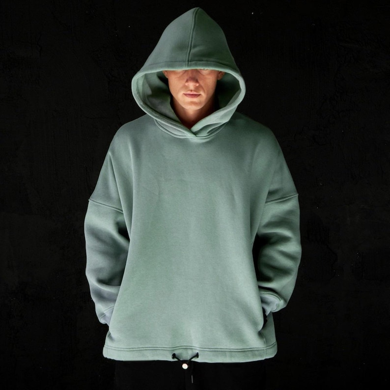 May include: A sage green hoodie with a drawstring at the waist. The hooded sweatshirt has a relaxed fit and long sleeves. The person in the image is wearing black pants. The background is black.
