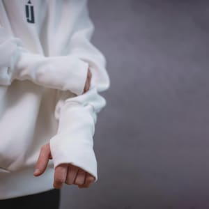 May include: A white hoodie with a black logo on the upper left chest. The sleeves have thumb holes. The person is wearing black pants. The background is a solid gray color.
