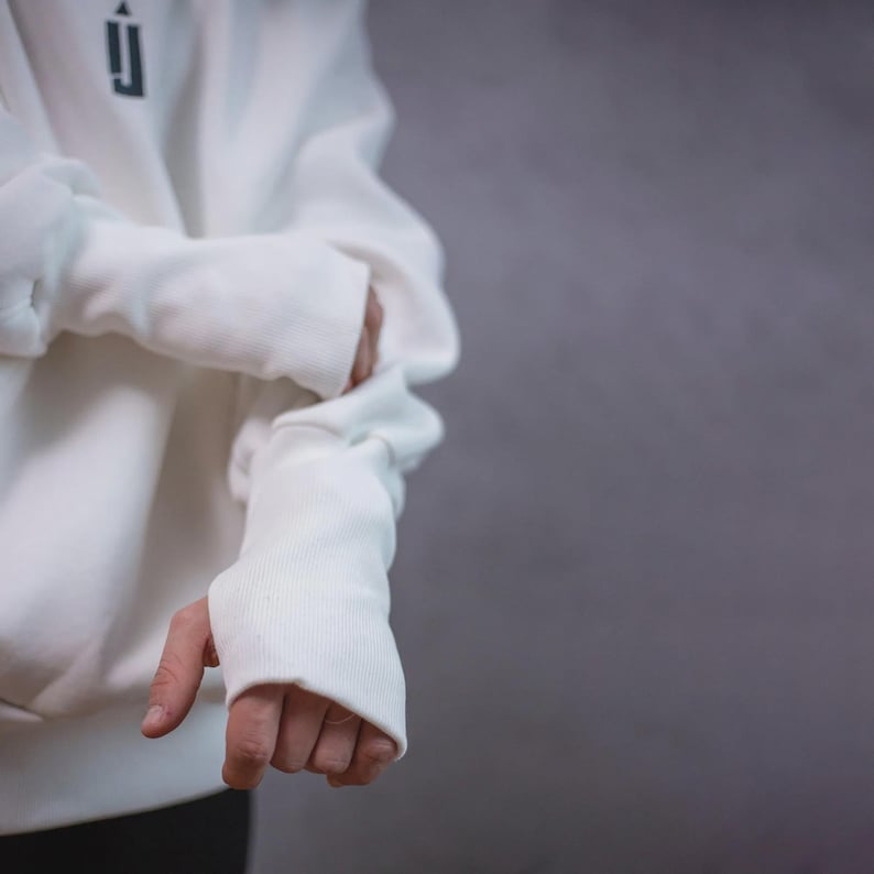 May include: A white hoodie with a black logo on the upper left chest. The sleeves have thumb holes. The person is wearing black pants. The background is a solid gray color.