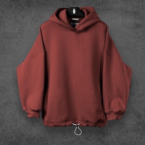 May include: A maroon hoodie with a drawstring at the waist and a hood. The hoodie is hanging on a black hanger against a gray background. The sleeves are long and the material appears to be a soft fabric.