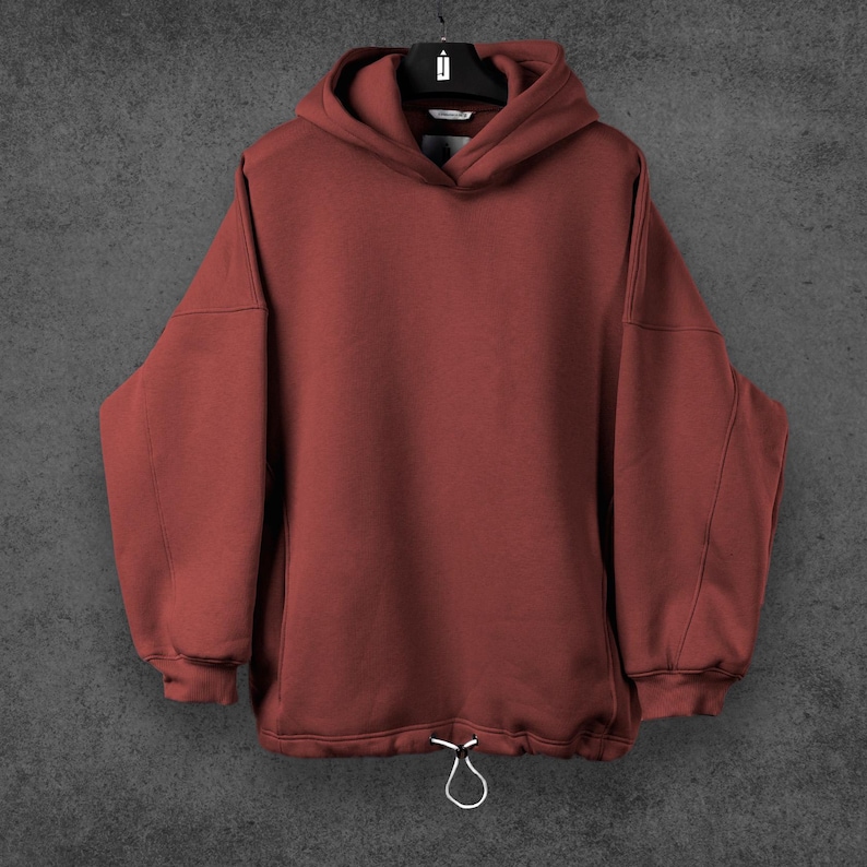 May include: A maroon hoodie with a drawstring at the waist and a hood. The hoodie is hanging on a black hanger against a gray background. The sleeves are long and the material appears to be a soft fabric.