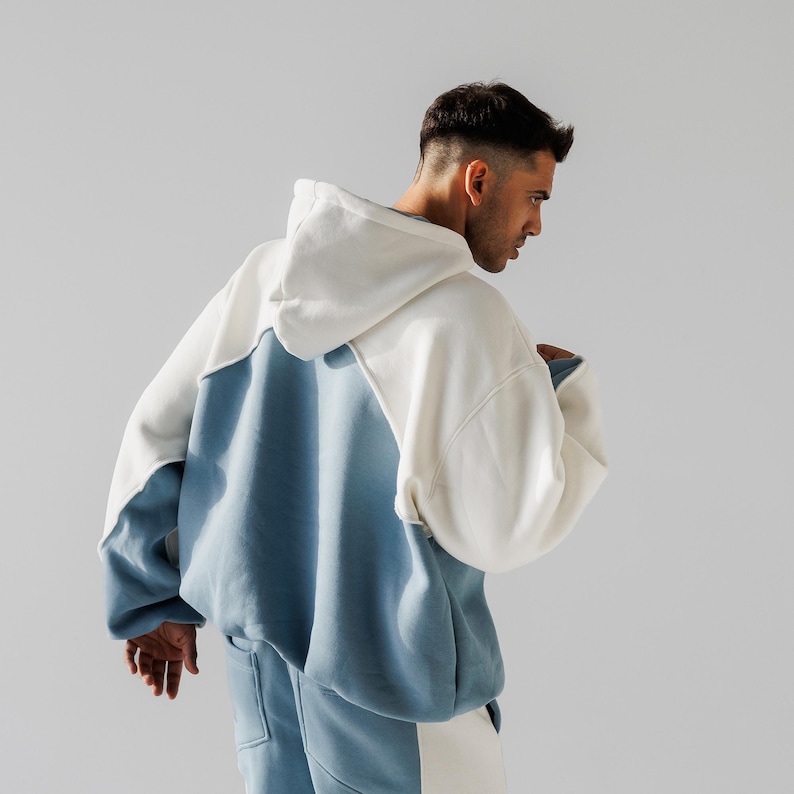 May include: A two-tone hoodie in white and blue. The hoodie features a large hood and a relaxed fit. The sleeves and lower portion of the hoodie are blue, while the upper portion and hood are white. The hoodie is part of a matching set.
