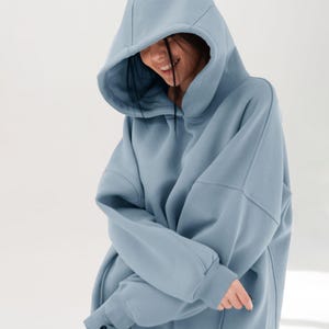 May include: A light blue hoodie with a large hood. The hoodie has a relaxed fit and long sleeves. The person wearing the hoodie is smiling. The hoodie is made of a soft material.