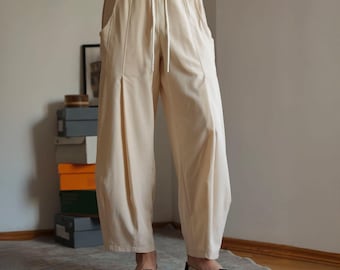 May include: Cream-colored wide-leg pants with a drawstring waist and side pockets. The pants are made of a lightweight fabric and are paired with black slip-on shoes. The model is wearing a white t-shirt.