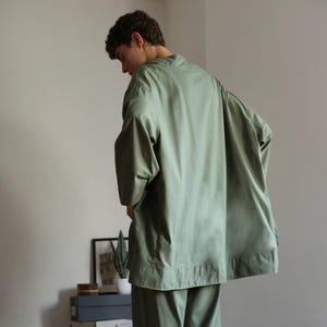 May include: A light olive green, oversized shirt and matching trousers. The shirt has wide sleeves and a relaxed fit. The outfit is made of a soft, lightweight fabric. The model is wearing the outfit.